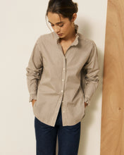 Load image into Gallery viewer, A woman wearing a beige and white vertically striped button-down shirt with dark blue jeans, standing against a neutral cream wall next to a wooden panel.
