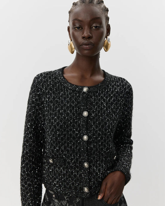 Woman wearing a black textured jacket with silver buttons on a light gray background black glitter cardigan women's fashion aprilmae chiswick