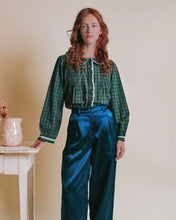 Load image into Gallery viewer, A woman with red hair stands indoors wearing a dark green plaid long-sleeve blouse with white trim and high-waisted teal satin trousers. She is posed next to a small white table with a pink ceramic vase against a plain beige wall.
